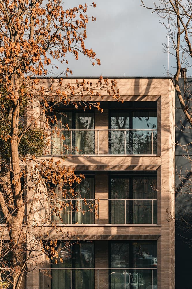 Nowoczesny, trzypiętrowy budynek Remedium z dużymi szklanymi oknami i balkonami, częściowo zasłonięty przez bezlistne drzewa z brązowymi liśćmi, pod zachmurzonym niebem.