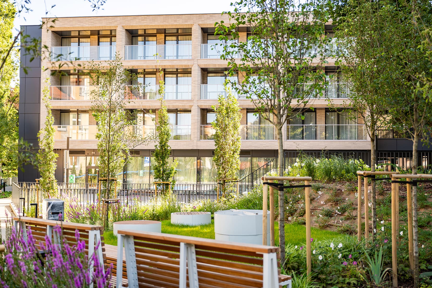 Nowoczesny trzypiętrowy budynek z dużymi przeszklonymi balkonami wychodzi na ogród krajobrazowy z ławkami, młodymi drzewami i kwitnącymi kwiatami na pierwszym planie.