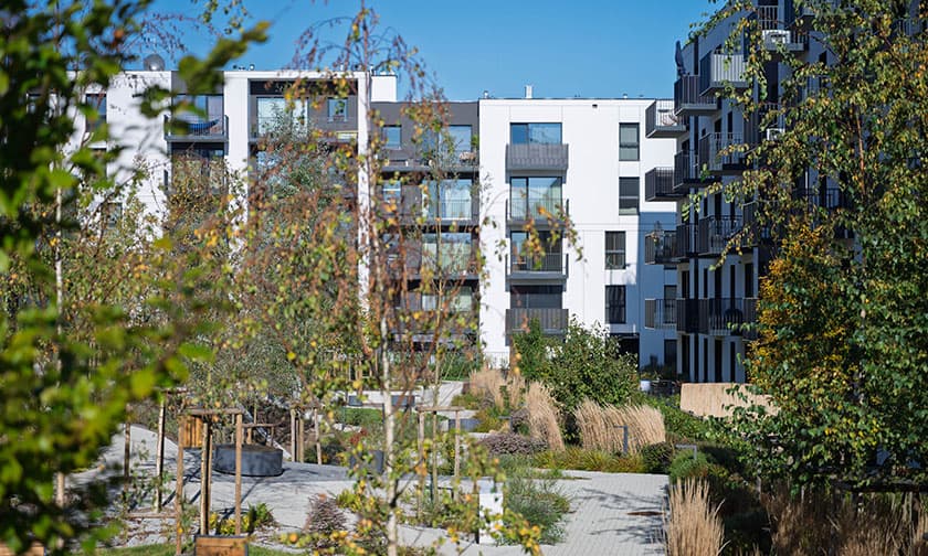 Nowoczesne apartamentowce z balkonami, otoczone zielenią, drzewami i ścieżkami w słoneczny dzień. Skorzystaj z rabatów do 22 000 zł na nowe mieszkania w tej wyjątkowej lokalizacji.